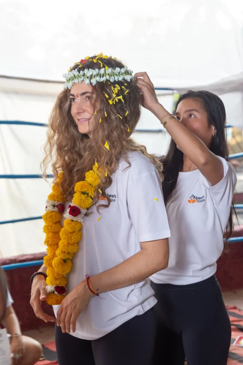 Student in an Alpesh Yoga t-shirt adjusting the jasmine flower crown of a garlanded classmate during the closing ceremony