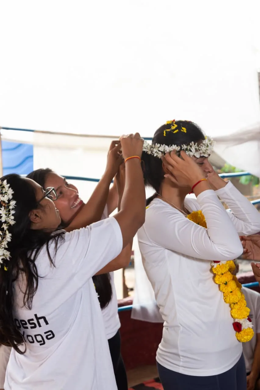 Laughing students fastening a jasmine flower crown onto a classmate during the closing ceremony of their yoga teacher training