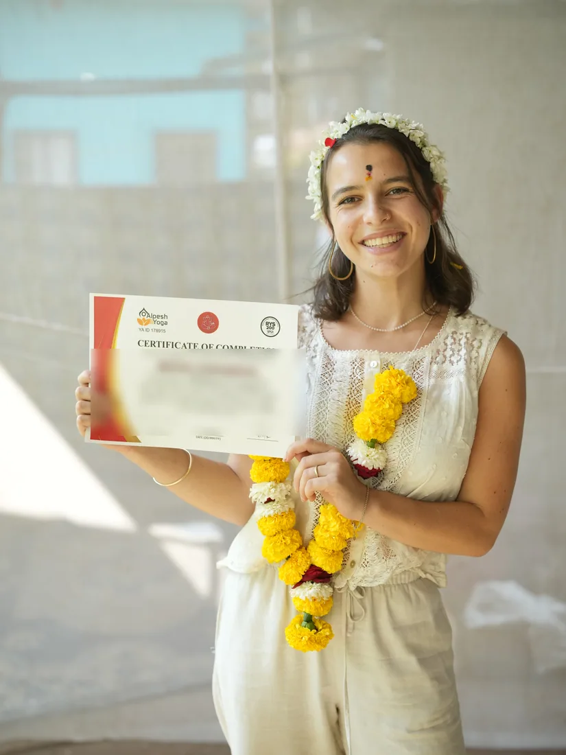 Smiling Alpesh Yoga graduate holding her 200-hour yoga teacher training certificate of completion, wearing a marigold garland and flower crown