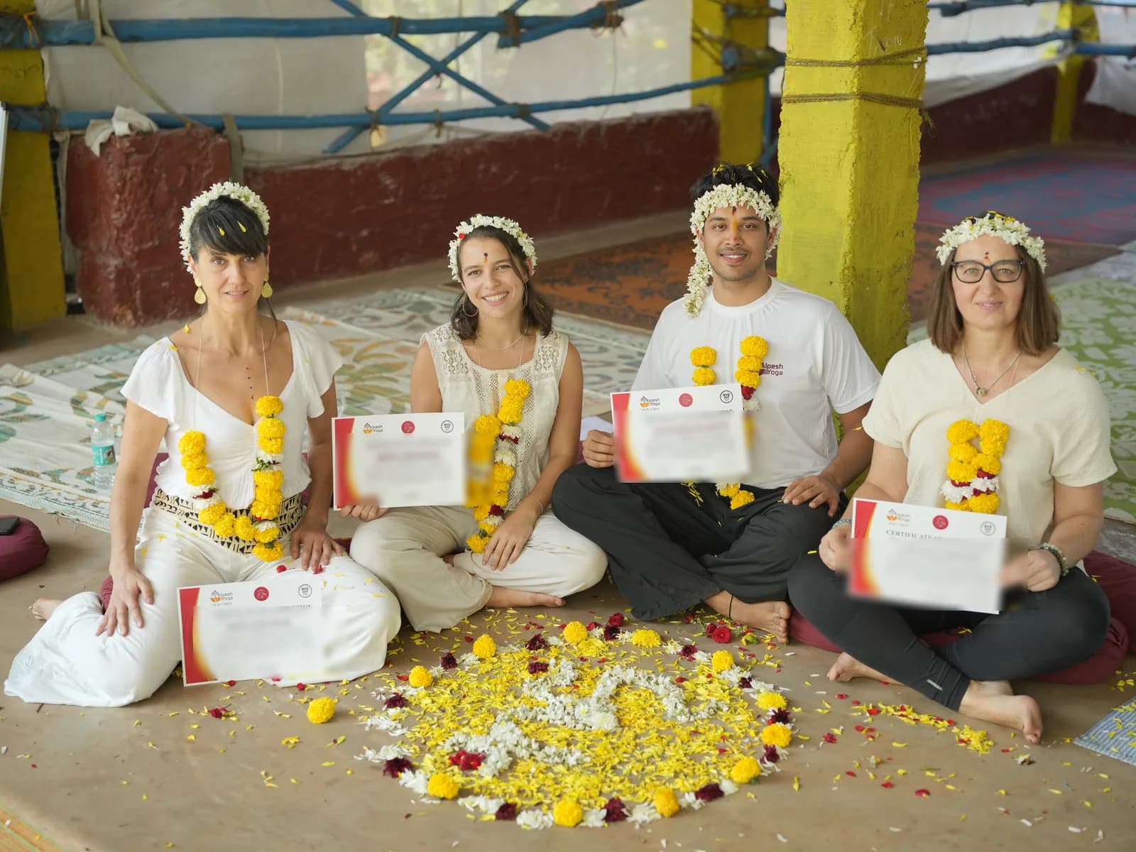Four Alpesh Yoga graduates seated around a marigold flower mandala, each holding their 200-hour yoga teacher training certificate of completion