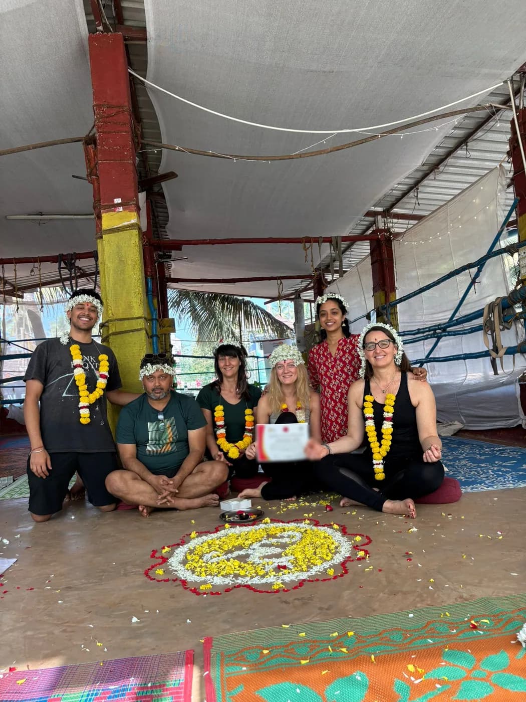 Alpesh Yoga graduating batch with flower crowns and marigold garlands gathered around a flower mandala with a certificate of completion