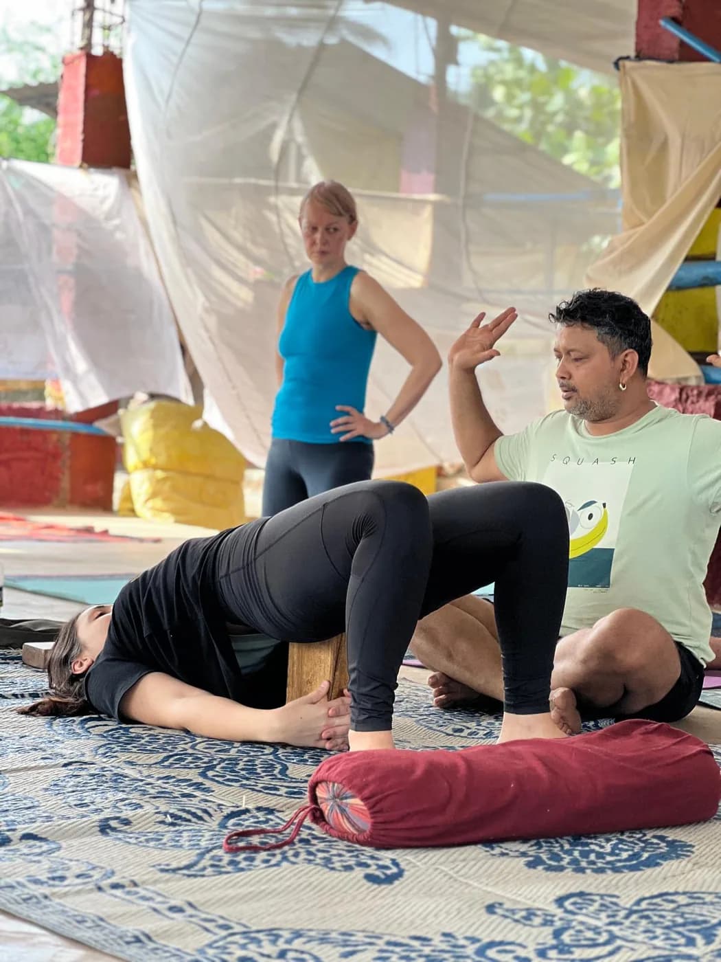 Teacher guiding a student through a block-supported bridge pose while the class observes