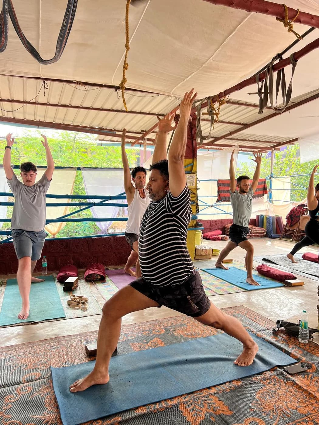 Yoga teacher training class practising Warrior I together in the open-air shala at Arambol, Goa