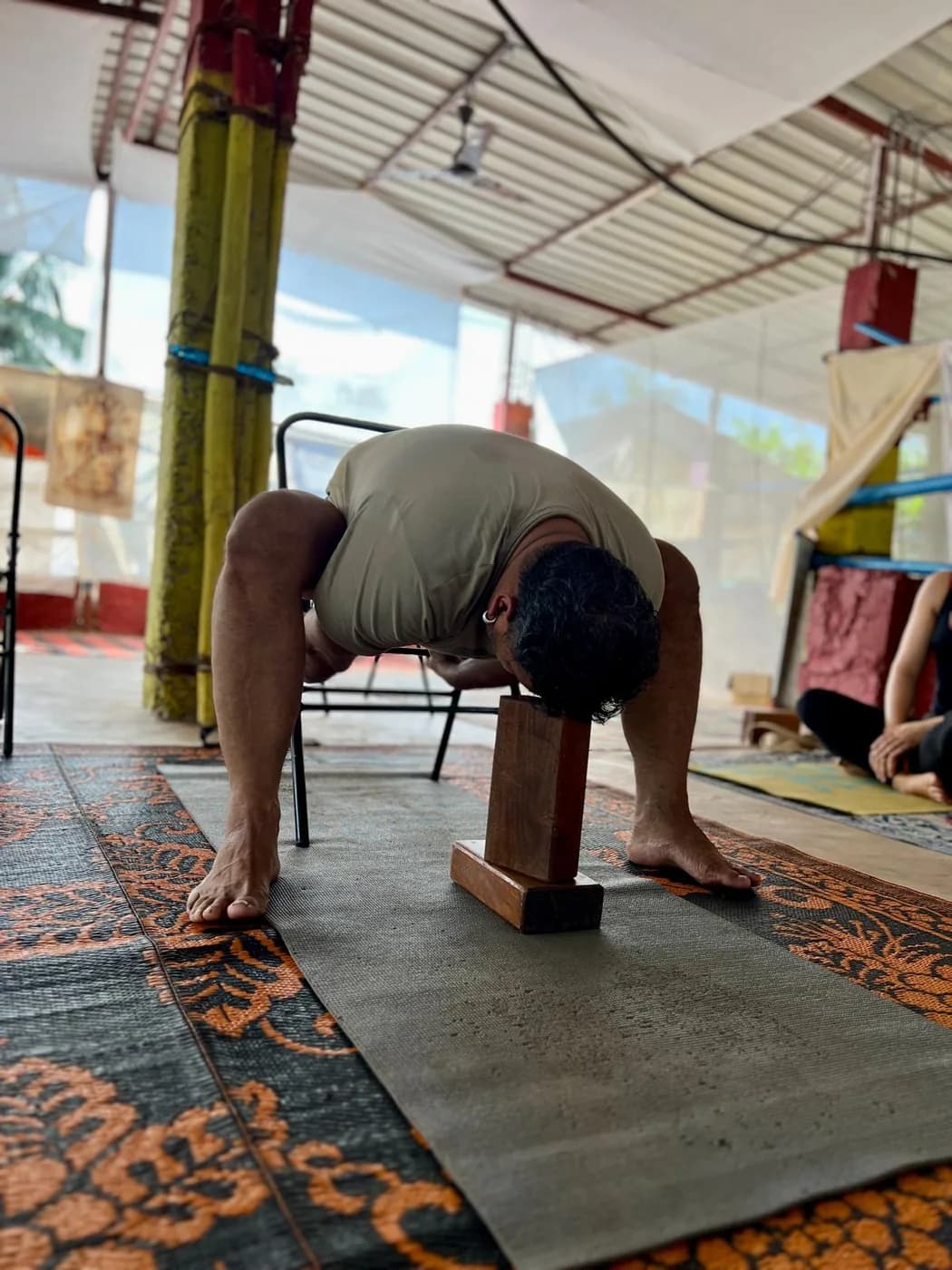 Teacher demonstrating a wide-legged standing forward fold with his head supported on wooden blocks while students watch