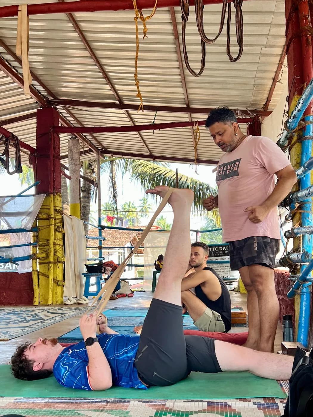 Student lying on his back extending one leg with a yoga belt while the teacher guides the alignment